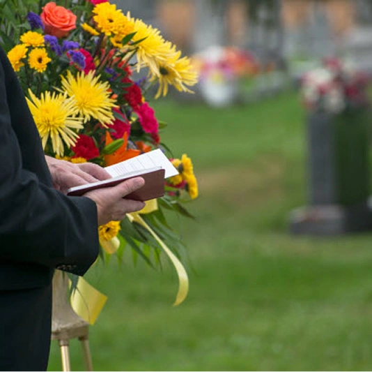 Person in a dark suit holding a bright bouquet of flowers and a small book at a cemetery funeral scene